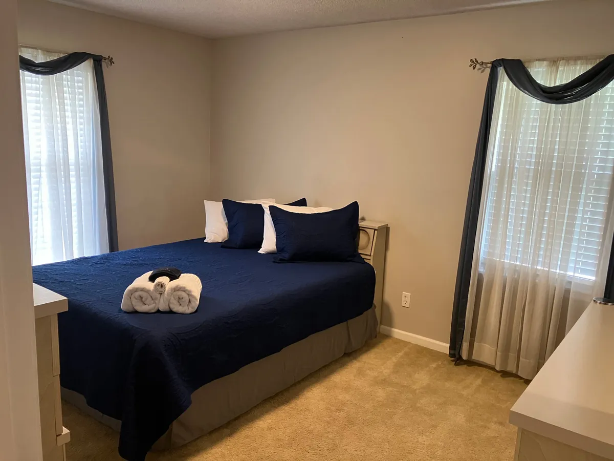 A serene bedroom with a neatly made bed, decorative towels, and large windows letting in natural light. The calm palette and soft textures contribute to a relaxing environment designed for restful nights.