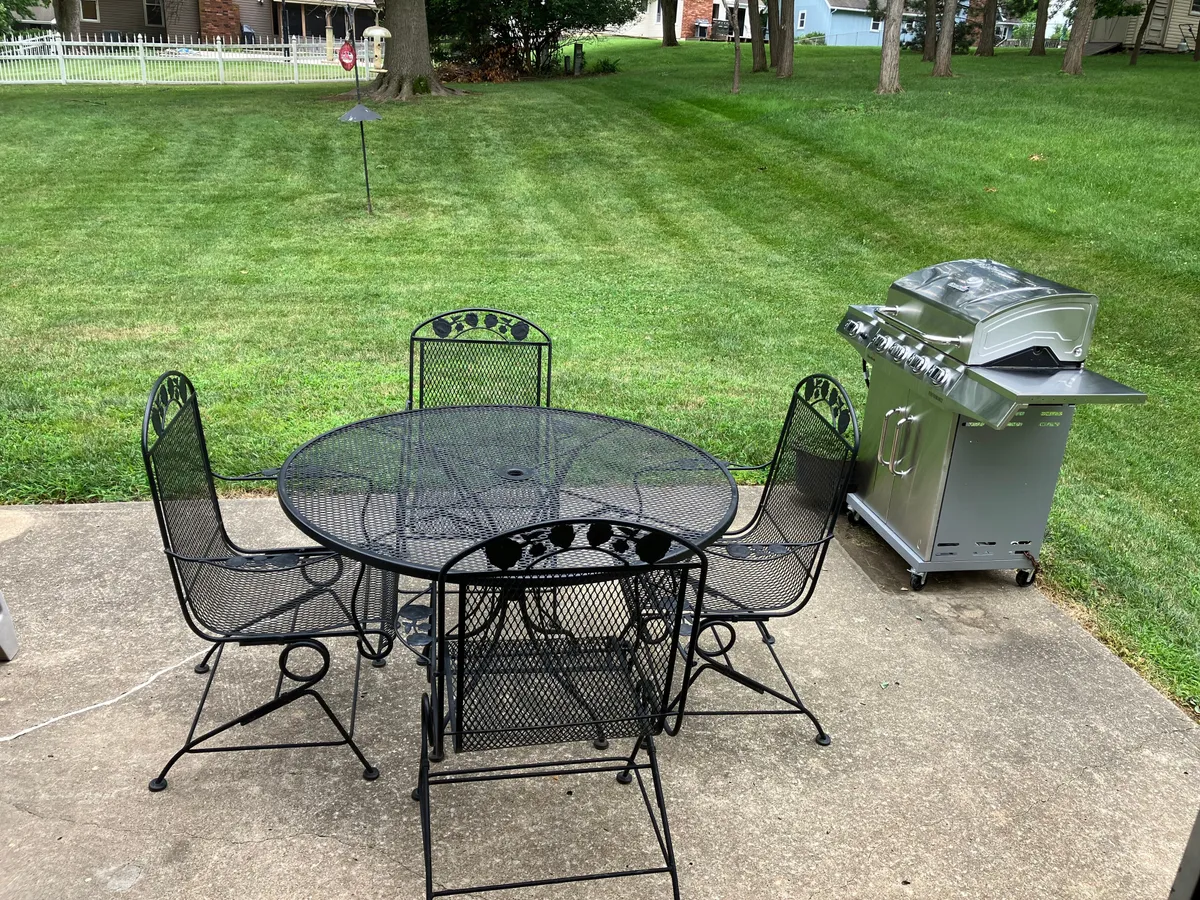This image features a well-maintained outdoor patio space with a black metal table, four matching chairs, a stainless steel grill, and a lush, manicured lawn. The scene conveys a welcoming and versatile area perfect for dining or relaxing outdoors.
