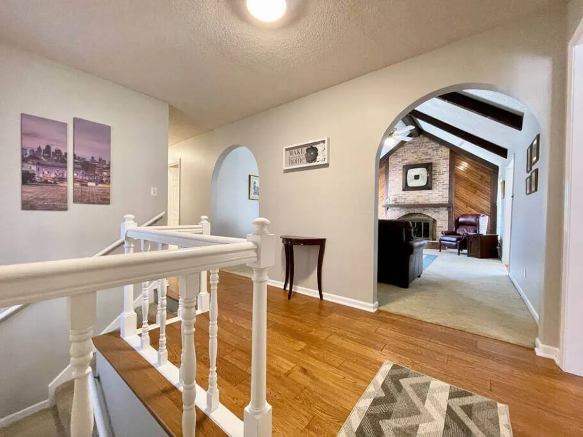 A bright entryway reveals elegant staircase railings, arched doorways, striking artwork, and a glimpse of a cozy fireplace in the adjacent living area. The space feels open and welcoming, suggesting ease of movement from public to private areas within the home.