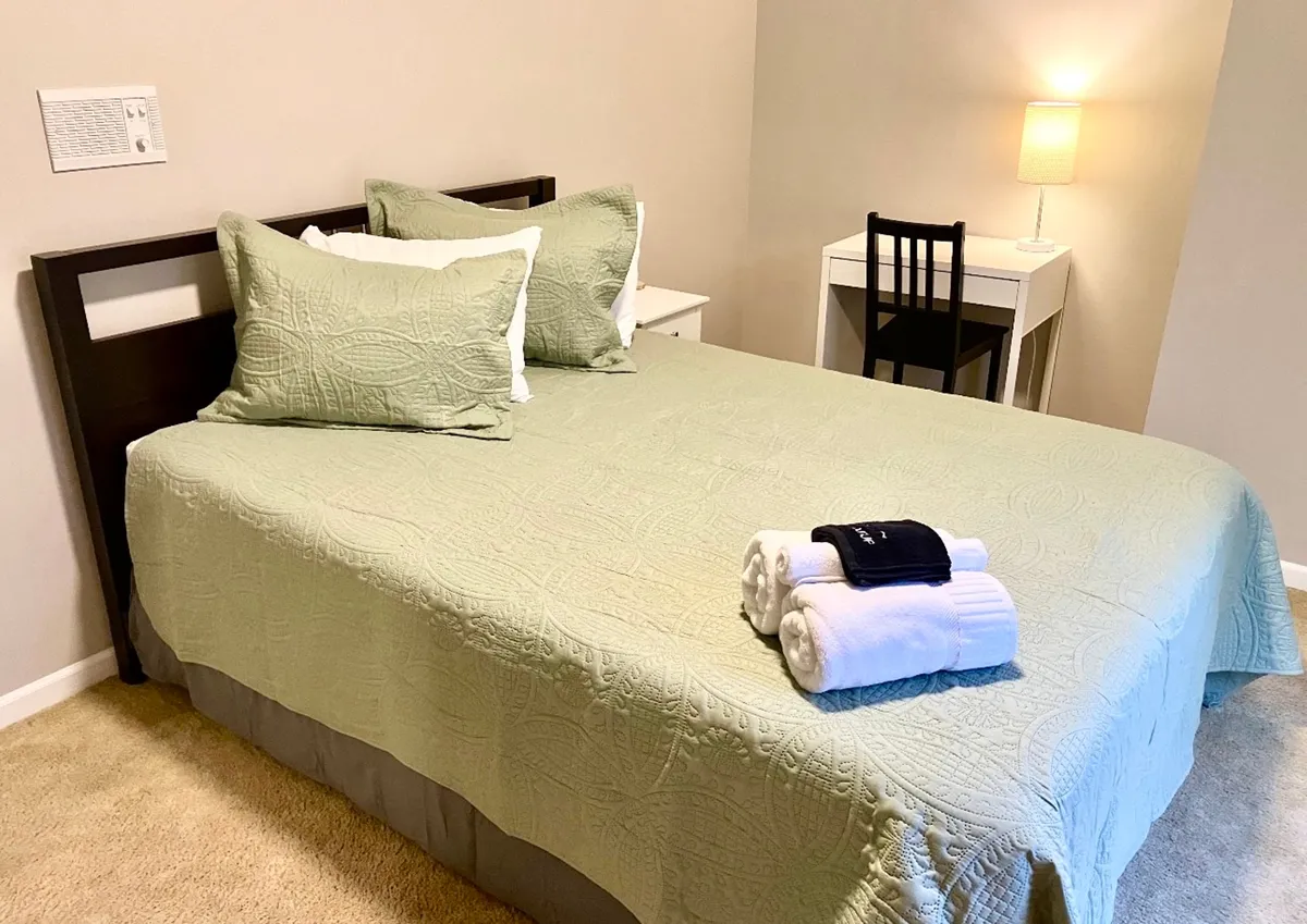 A serene bedroom with a bed dressed in a green quilt and decorative pillows, complemented by white towels placed at the foot. A small work desk with a lamp sits nearby, providing a quiet nook for productivity. The overall mood is cozy and relaxing, ideal for both rest and work.