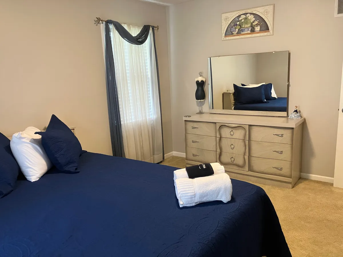 A calm, comfortable bedroom featuring a stylish bed with navy blue bedding, a dressing table with mirror, and tasteful wall decor. The room is bright and inviting, ideal for relaxing, getting ready, or enjoying a quiet moment alone.